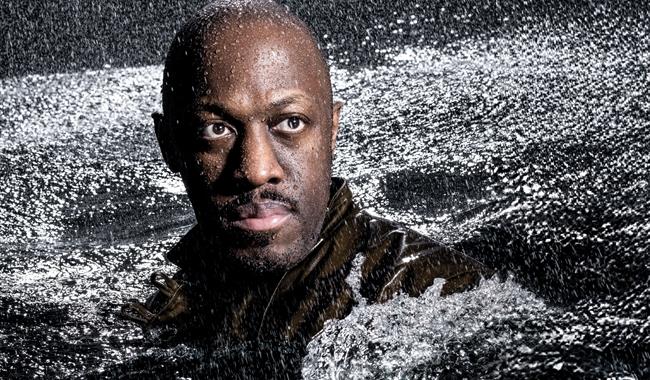A man's head and shoulders rise from a sea of rough, churning water; rain is pouring down on him. He stares into the distance, his face illuminated by