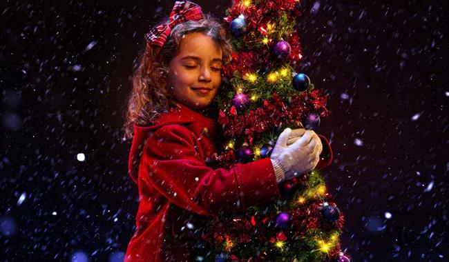 A young girl in a red winter coat hugs a small Christmas tree. She is smiling contentedly with her eyes closed, and has a red tartan bow in her hair.