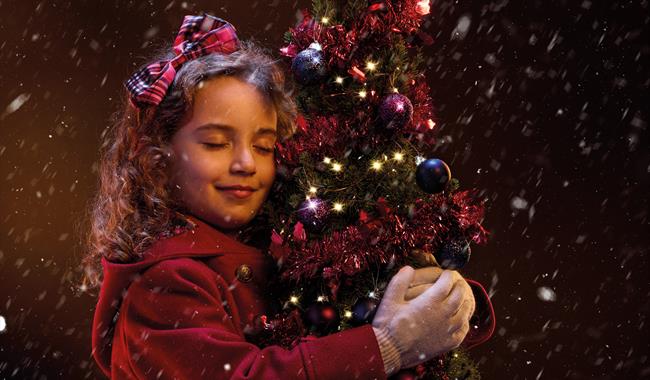 A young girl in a red winter coat hugs a small Christmas tree. She is smiling contentedly with her eyes closed, and has a red tartan bow in her hair.