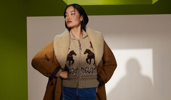 Lady dressed in a layered outdit with coat, zip up jumper and pair of jeans against a olive green background