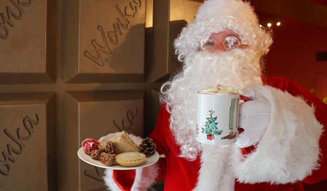 Santa stood in front of some giant chocolate bar doors holing a cup of hot chocolate and a plate of sweet treats, including a mince pie