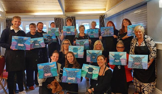Painting party group holding finished paintings on canvases of Highland cows