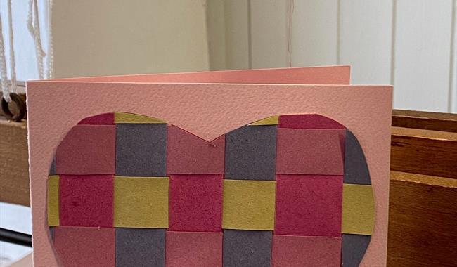 image of heart woven from paper on a card which is sitting on top of a wooden weaving loom