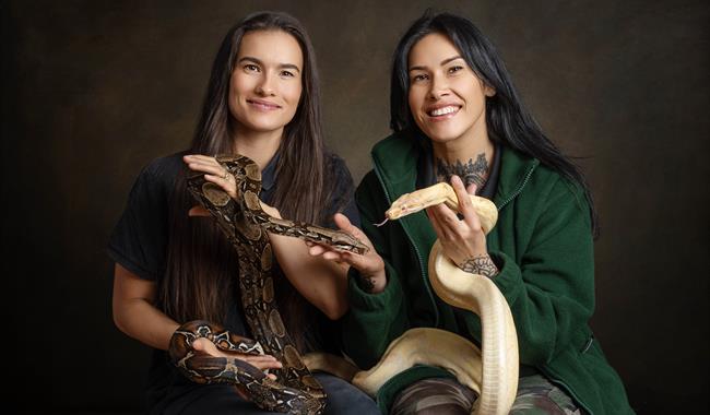 Two women sat against a dark background, holing a cream-coloured snake