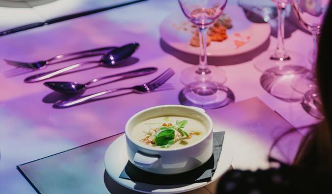 A dinner table, with a colourful print projected onto it. Soup in a bowl.