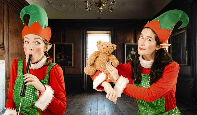 Two women dressed as Christmas elves inside Admiral's House. One is holding a Teddy Bear and the other one is playing the flute.