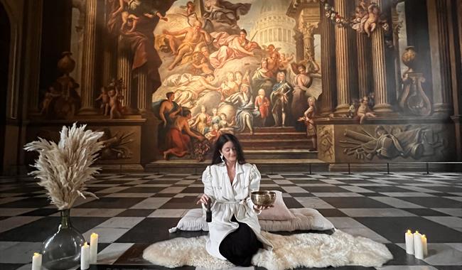 A person dressed in a white robe sits cross-legged on a white fur rug in an ornate hall with checkered floors. A large classical mural decorates the w