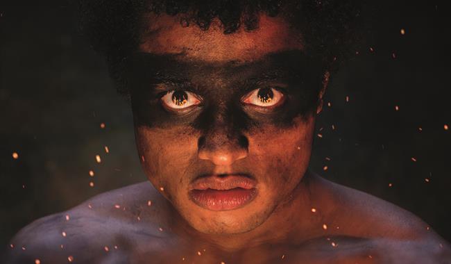 A boy stares brutally straight into the camera with a straight face. There is messy black dirt over his eyes, like a mask, and he is topless with dirt