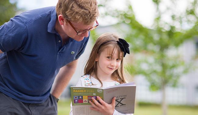A girl at Bletchley Park looks towards the camera as her dad tries to see the answers in the mission activity booklet she is completing.