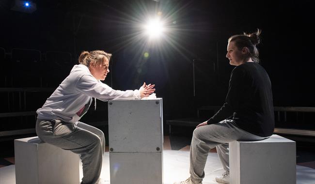 Two women sit on white blocks on a bright white stage; the background is in darkness but there's a central stage light glaring out front. The woman on