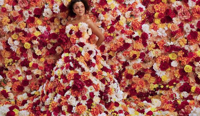 A young woman stands totally surrounded by flowers. They flood the background and cover her strapless, floor-length gown. The flowers are a mixture of