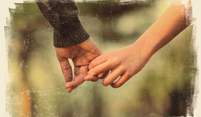 Two hands with two fingers linking, pulling away slightly. One has a bare arm and the other wears a khaki jumper. The background is dappled green and