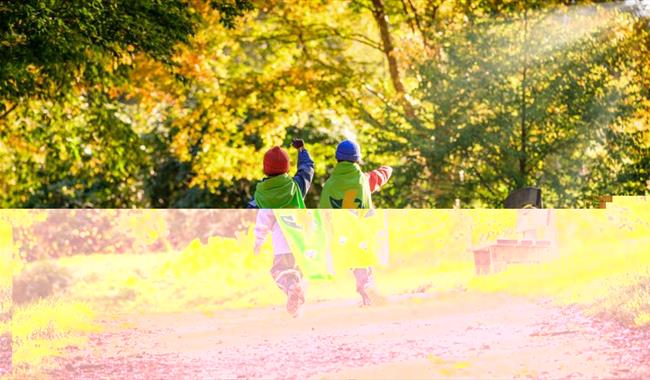 Two children run through autumnal woodland at Wakehurst wearing Nature Heroes capes