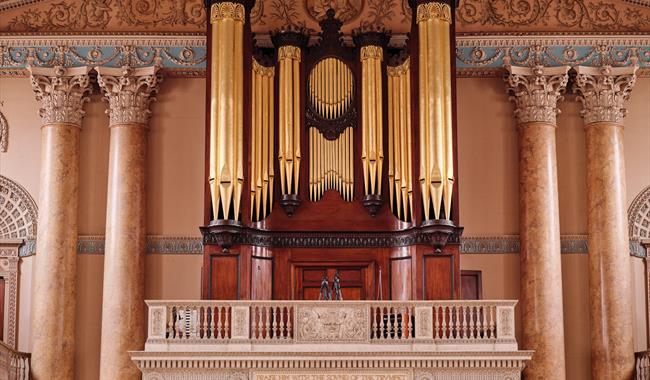 A grand pipe organ located in the Chapel of St Peter & St Paul featuring large golden pipes framed by dark wood and intricate architectural details. T