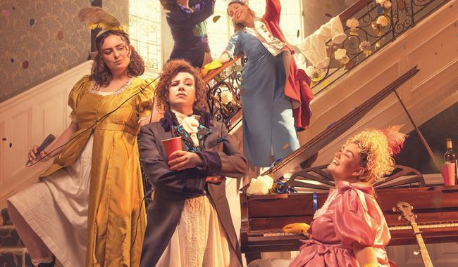 Five women dressed in Regency dresses but modern lace-up boots are having a party. In the foreground, a woman in a yellow satin dress and white pettic