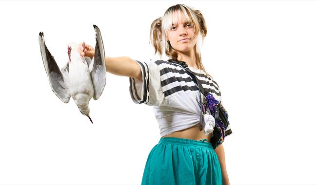 A young woman with blond hair tied up in bunches stares straight out front with a seagull thrust out in front of her, held in her right hand. She wear