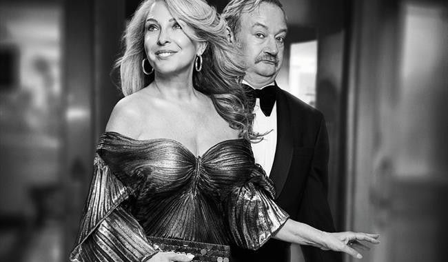 A black and white photo of a glamorous blonde woman in an off-the-shoulder pleated lurex dress and big gold hoop earrings, with a confident smile. Beh