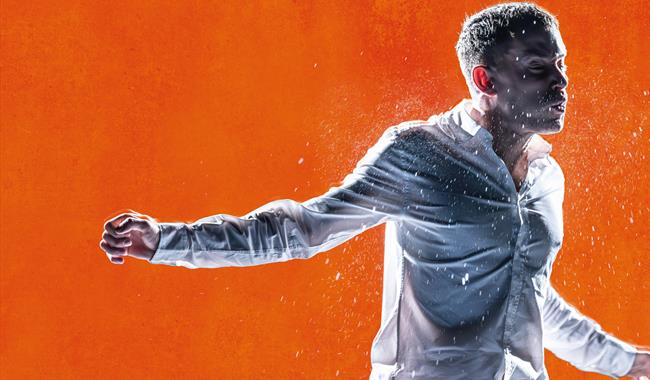Against a bright orange background, a young man is in motion, as though mid-fight. He wears a white shirt. He holds one arm out to the side and his fa