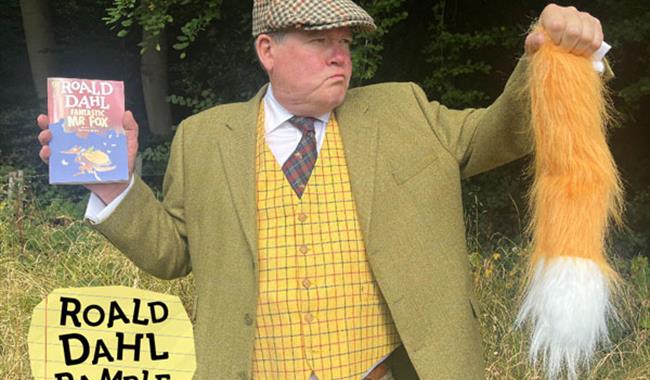 A man dressed as an old-fashioned farmer  is outside by some trees. He is hodling up a copy of the book Fantastic Mr Fox in one hand and a fox's tail