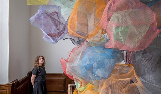 A women looks up a structure made from coloured mesh clouds that seem to float in the middle the stairwell of a wooden staircase.