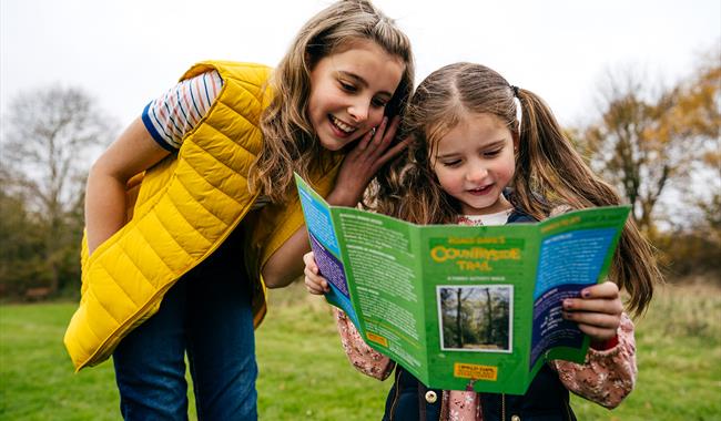 A child looking at a walk leaflet. She is stood in a green field, surrounded by trees