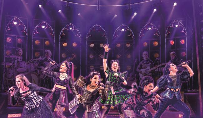 Six women in modern versions of Tudor outfits pose like a pop group, singing into handheld microphones and dancing in a variety of lively and energeti