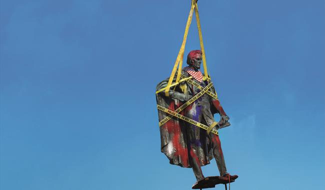 A traditional stone statue of a historic figure is suspended from an orange metal hook with yellow straps against a bright blue background. It is wrap