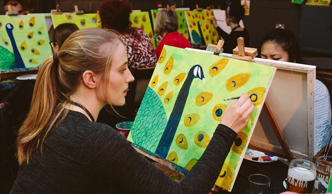 Person smiling, relaxed, sitting down in a group setting with a canvas on an easel with paintbrushes in a c up beside it