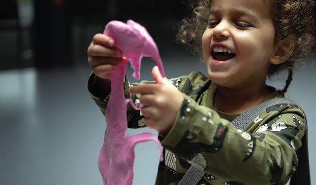 A young girl holding pink slime. The girl is looking at the slime with a big smile on her face.