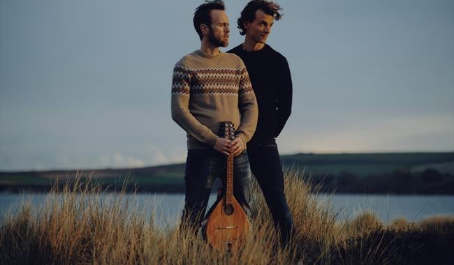 Two middle aged white men with dark hair and facial hair stand on the cliffs looking wistfully into the distance
