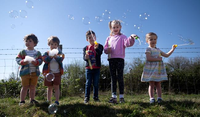 5 children stood outdoors with bubble wands and bubbles around them. The sky is bright blue in the background. 2 boys are holding foam in their hands.