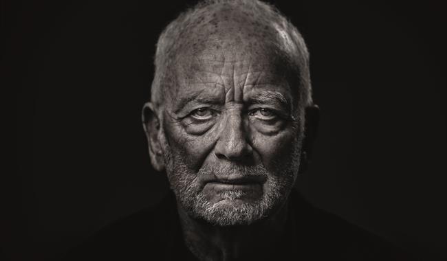 An elderly man (Ian McDiarmid) stares at the camera. His face is wrinkled and looks stern and dramatic, with shadows under his eyes and chin.  He is w
