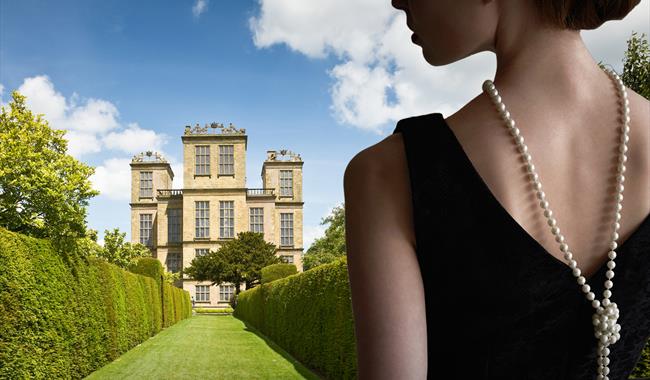 Blue sky with white clouds are the backdrop to a stateley home, with green manicured lawns stretching out in front, at the foreground of the image. To