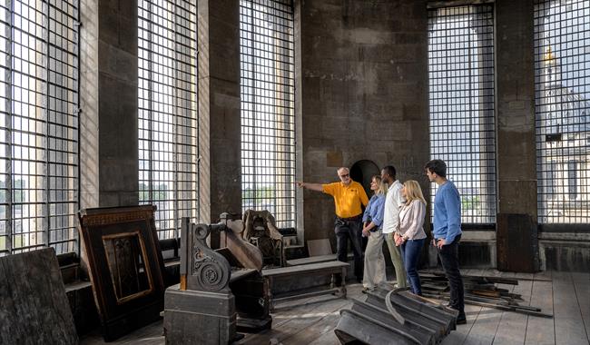 A group of five people, guided by a man in a yellow shirt, are standing inside a large room with tall, grid-patterned windows inside the Chapel Dome a