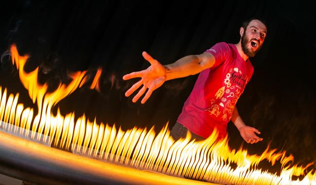 A man with a pink T-shirt on is standing in front of a large metal tube that has lots of holes in it. There is fire coming out of the holes and the ma