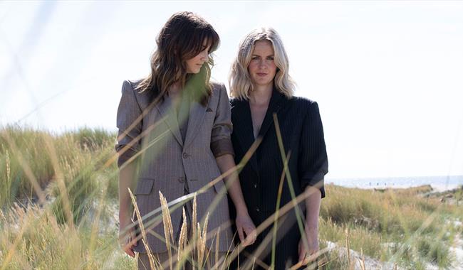 photograph of two women in grey and black suit jacket, one blonde and one brunette, standing on sand dunes amongst the grasses.