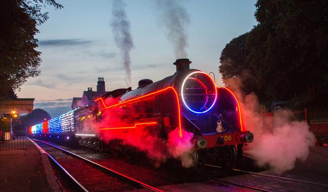 A steam locomotive travelling along a track with neon and fairy lights on the train.