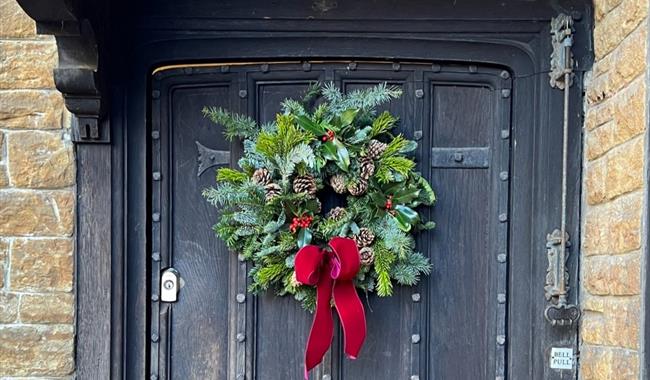 Photograph of a Christmas wreath made from fir branches with pine cones, berries and a red ribbon tied to it as decoration.