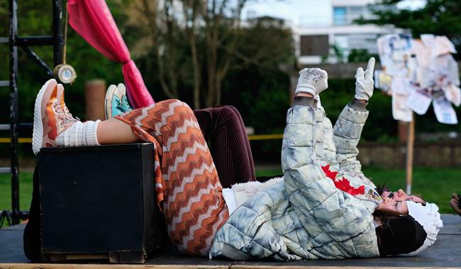 A person dressed in colourful clothing reclines on their back with legs propped up on a black box, pointing upward. The background includes trees, a b