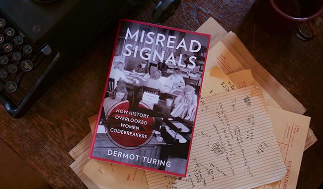A book called 'Misread Signals' by Dermot Turing resting on a wooden desk with various bits of paper with codebreaking on