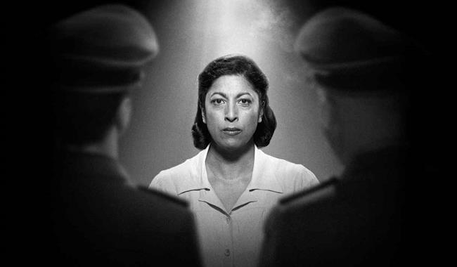 In a black and white photograph, a woman with dark shoulder-length hair stares resolutely at the camera, wearing a white top. A light illuminates her
