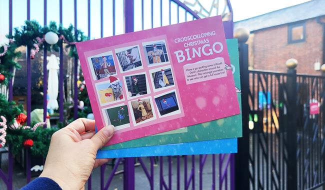 A hand holding a fan of bingo cards. In the bacground you can see some purple gates with a large Christmas wreath on them