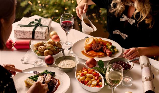 Guests enjoying a festive Christmas dinner with roast potatoes, seasonal vegetables, wine and all the trimmings beside a twinkling Christmas tree.