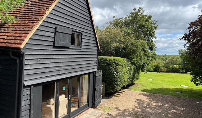 Black cladded barn with wide terrace doors. The view is a green field with trees in the far distance.