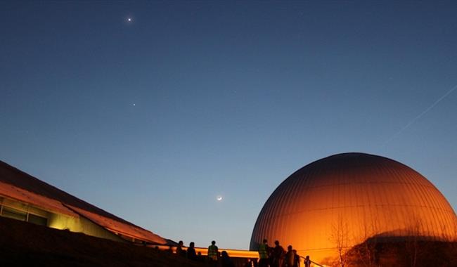 The Planetarium illuminated orange. Moon and stars in the sky next to the dome.