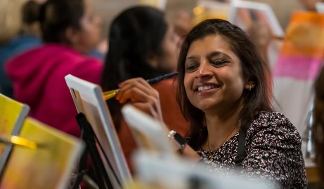 Person smiling, relaxed, sitting down, painting in a group setting with a canvas on an easel with paintbrushes