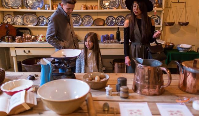 Three visitors stand around a wooden table in a historic kitchen or still room, surrounded by traditional cookware and kitchen tools at Uppark house a