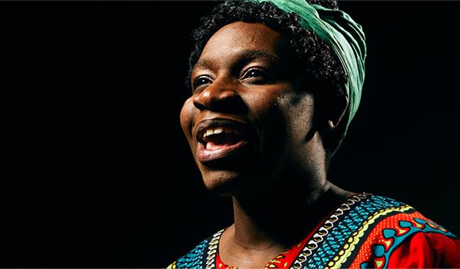 Against a black background a woman in a red, blue, black, green and yellow patterned top, in an African print, with a green hair-wrap, is looking off