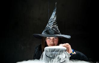 A young girl dressed as a witch with a cauldron bubbling over in front of her.