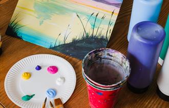 Painting class set up on a table including a tabletop easel with a finished landscape painting on canvas with red cup with paintbrushes neatly scatter